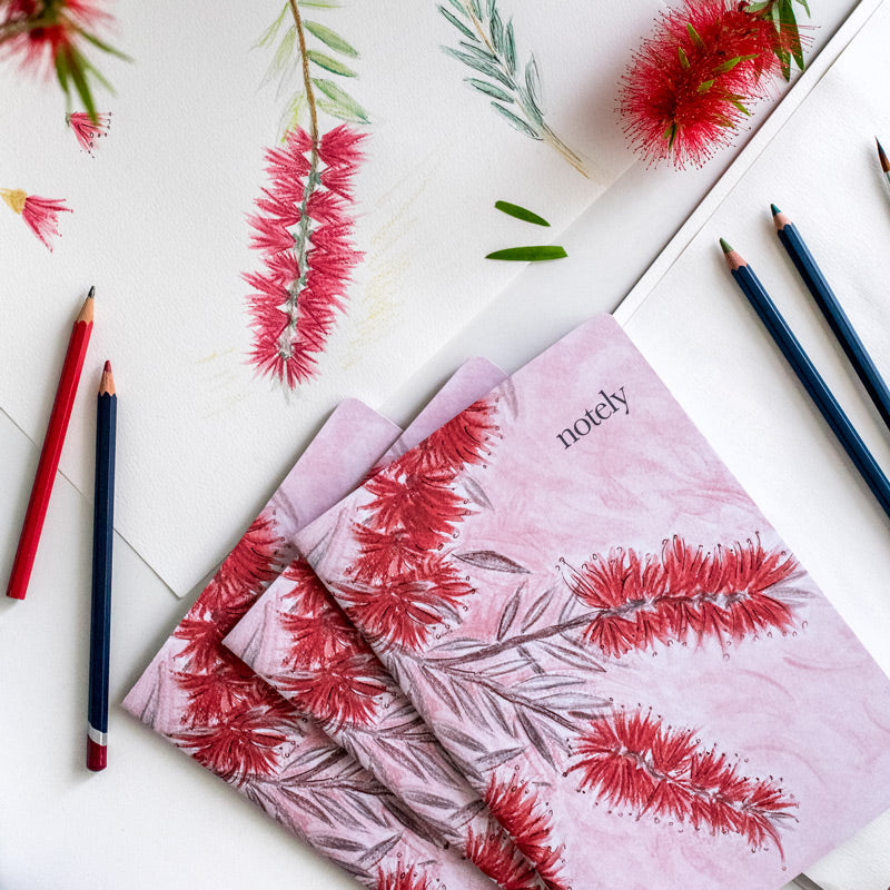 Flat lay of Notely Bottleblush notebooks on desk with artwork, bottlebrush flowers and pencils.